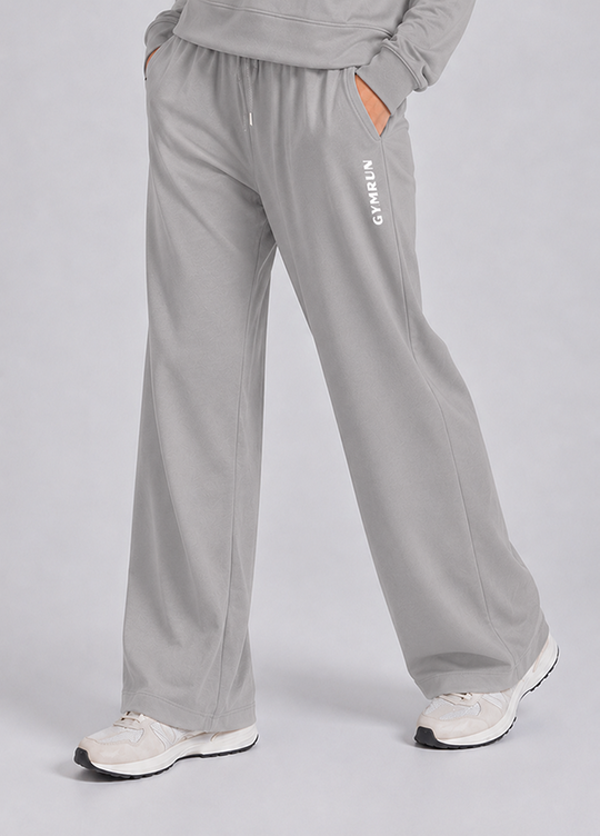A person wearing light gray wide-leg sweatpants with a white vertical 'GYMRUN' logo on the left leg, paired with white sneakers, shown from the waist down against a light gray studio background.