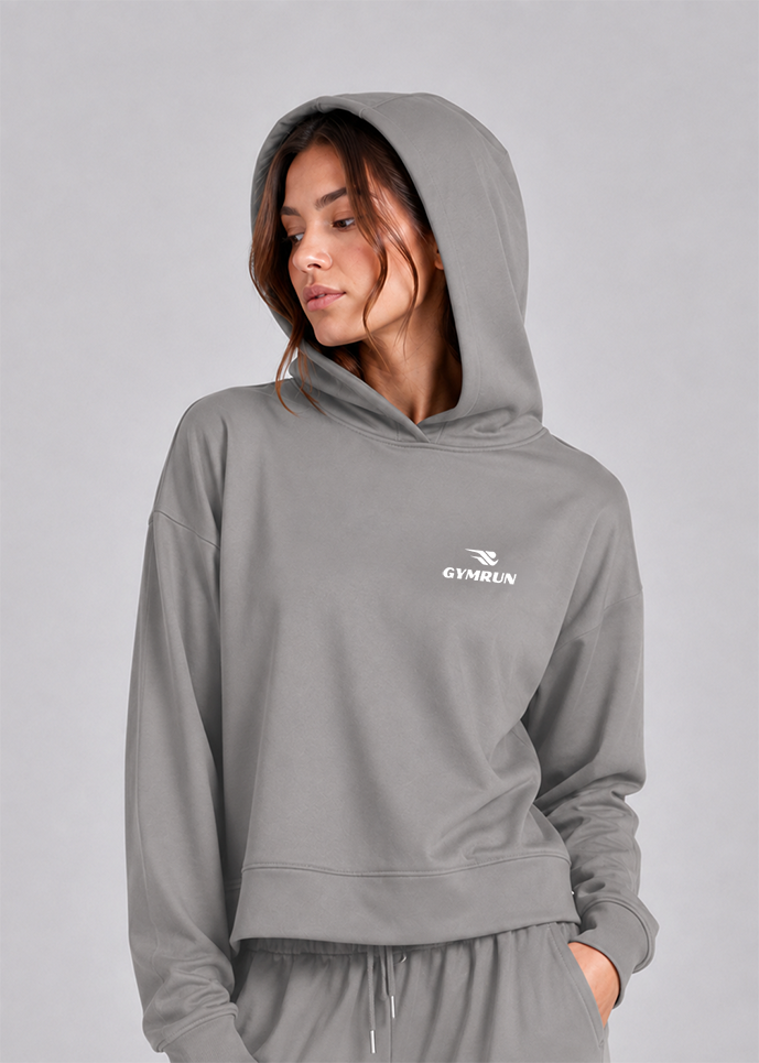 A woman wearing a gray cropped hoodie with a white GYHRUN logo on the left chest, paired with matching gray sweatpants, set against a solid gray studio background.