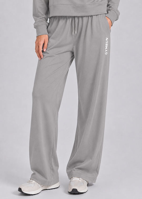 A person wearing light gray wide-leg sweatpants with a drawstring waist and white sneakers, shown from the chest down against a plain light gray studio background. The pants have vertical white 'GYMRUN' text on the left leg.