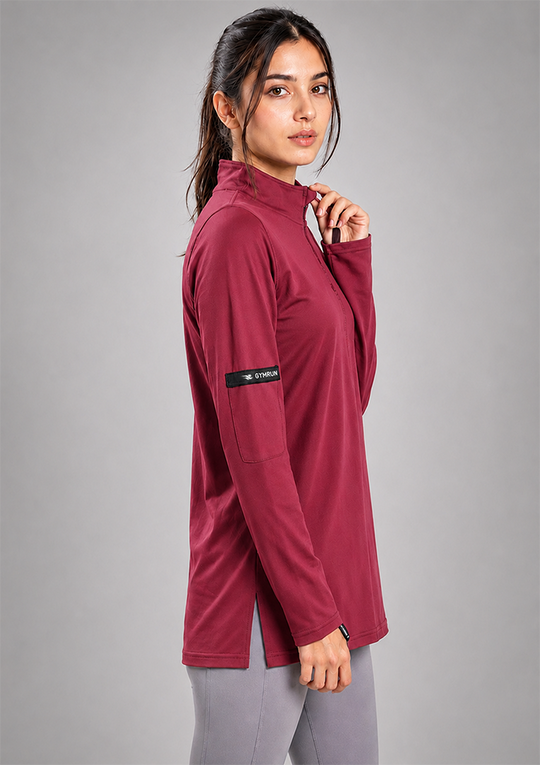 A woman modeling a long-sleeve athletic quarter-zip top in deep burgundy, paired with gray leggings, against a solid gray studio background.