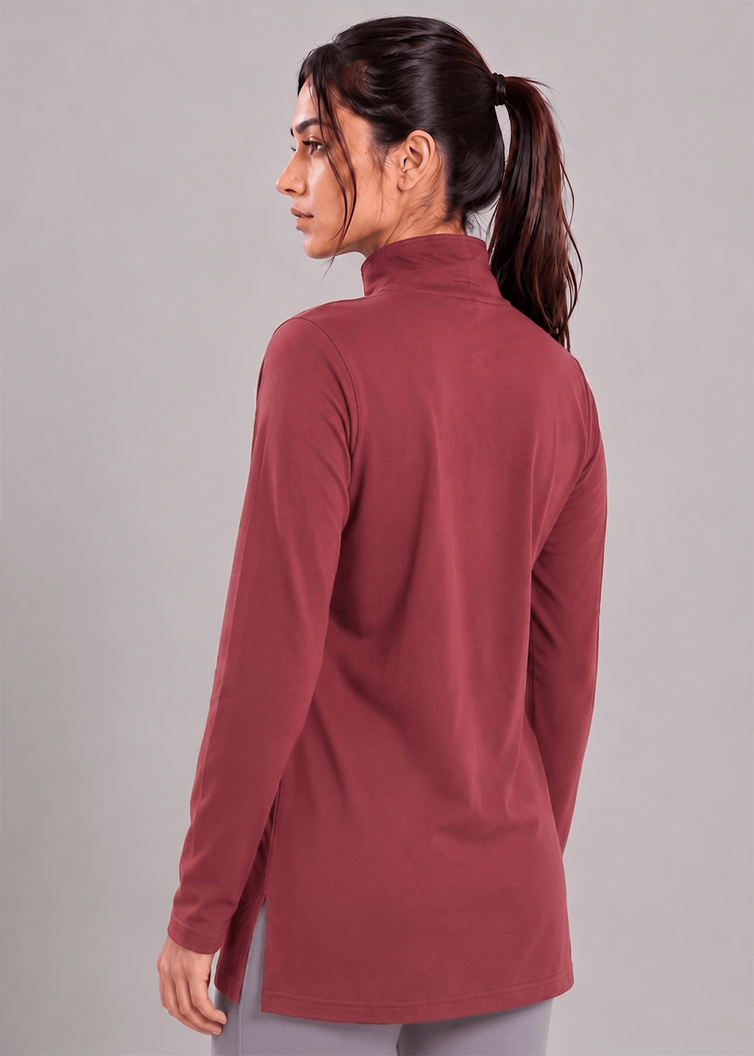 A woman with her hair in a ponytail is shown from the back wearing a long-sleeve mock-neck athletic top in a dusty rose or mauve color, paired with gray leggings, against a solid gray studio background.