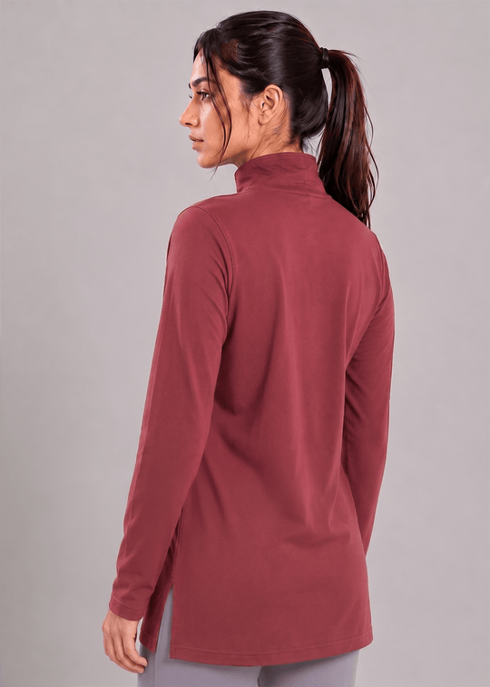 A woman with her hair in a ponytail is shown from the back wearing a long-sleeve mock-neck athletic top in a dusty rose or mauve color, paired with gray leggings, against a solid gray studio background.