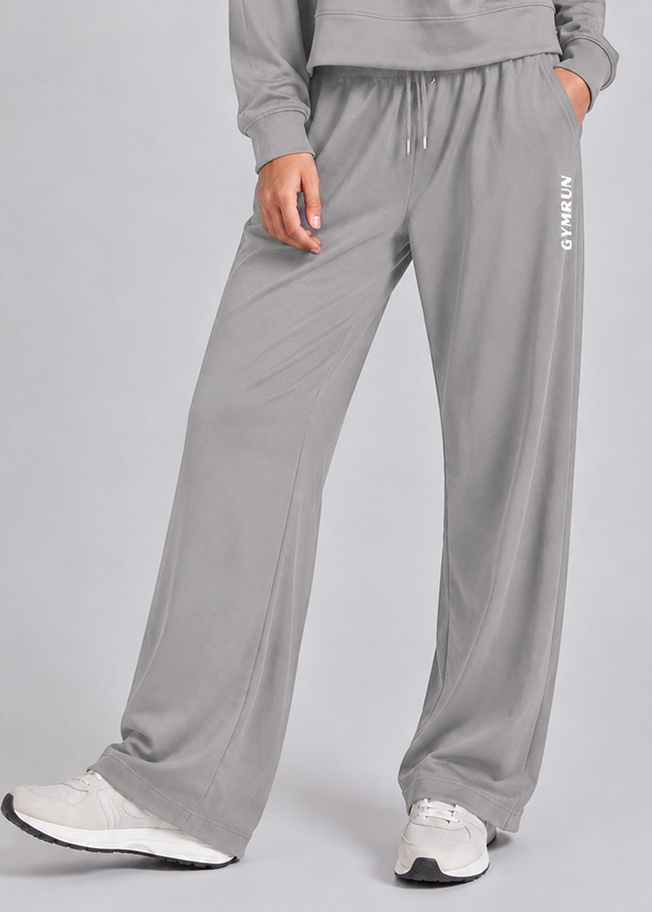 A person wearing light gray wide-leg sweatpants with a drawstring waist and white sneakers, shown from the torso down against a light gray studio background. The pants have vertical white 'GYMRUN' text on the left leg.