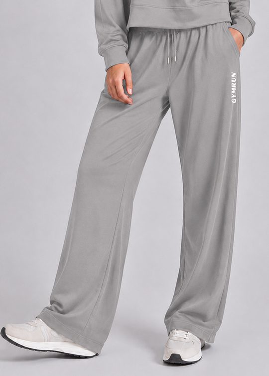 A person wearing light gray wide-leg sweatpants with a drawstring waist and white sneakers, shown from the torso down against a light gray studio background. The pants have vertical white 'GYMRUN' text on the left leg.
