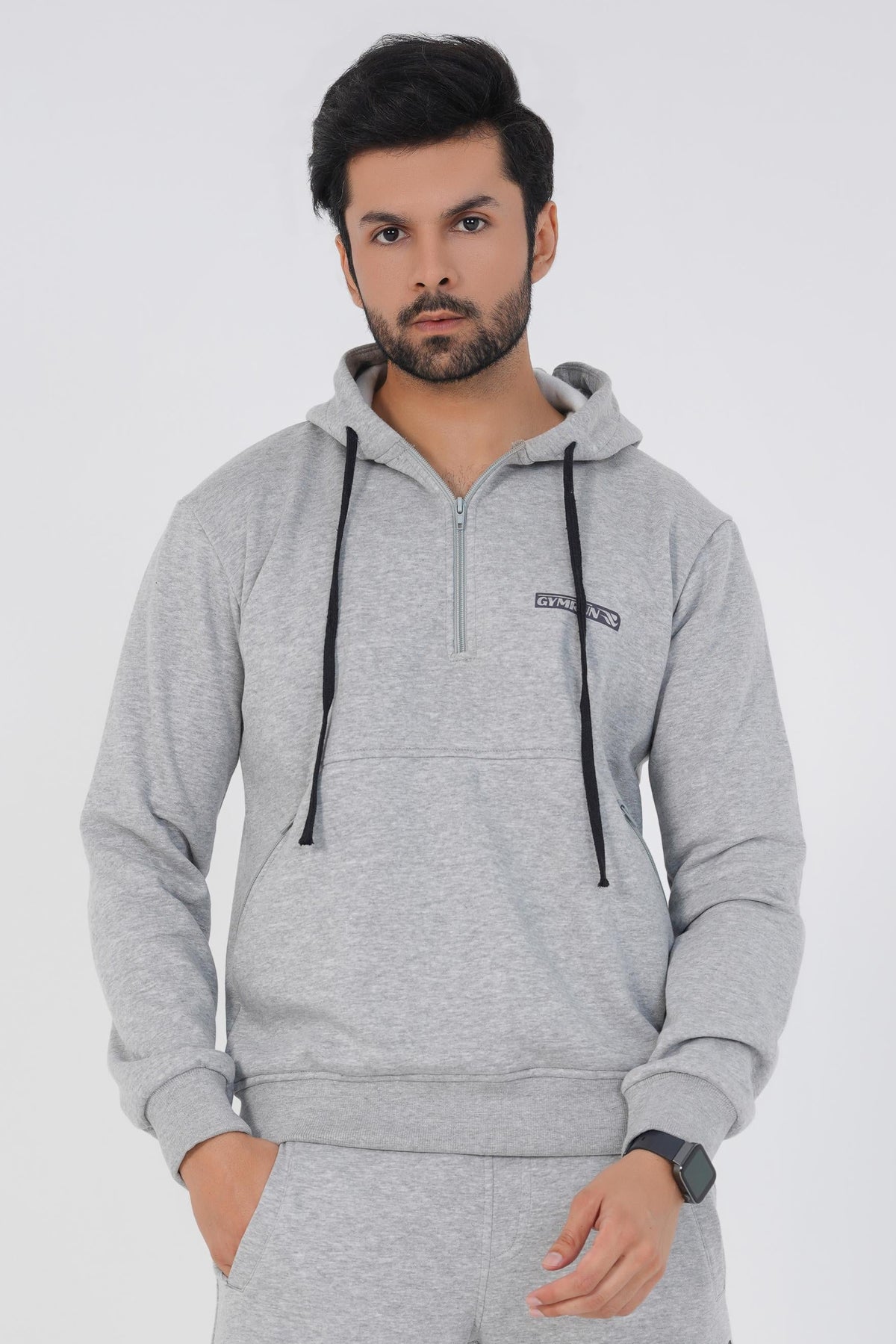 A man wearing a heather grey hoodie with a three-piece hood, adjustable drawcord, and a front pouch pocket with zipper. He is also wearing matching grey pants and white sneakers.