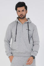 A man wearing a heather grey hoodie with a three-piece hood, adjustable drawcord, and a front pouch pocket with zipper. He is also wearing matching grey pants and white sneakers.
